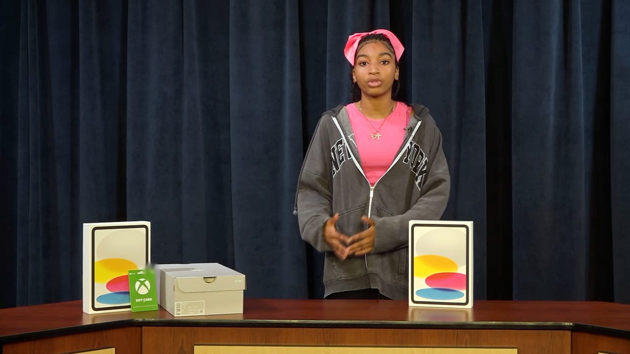 A teenage student in a gray zip hoodie and a pink headband stands behind a wooden desk in front of a navy stage curtain, speaking toward the camera. Two tablet boxes, a shoe box, and a gift card are lined up on the desk.